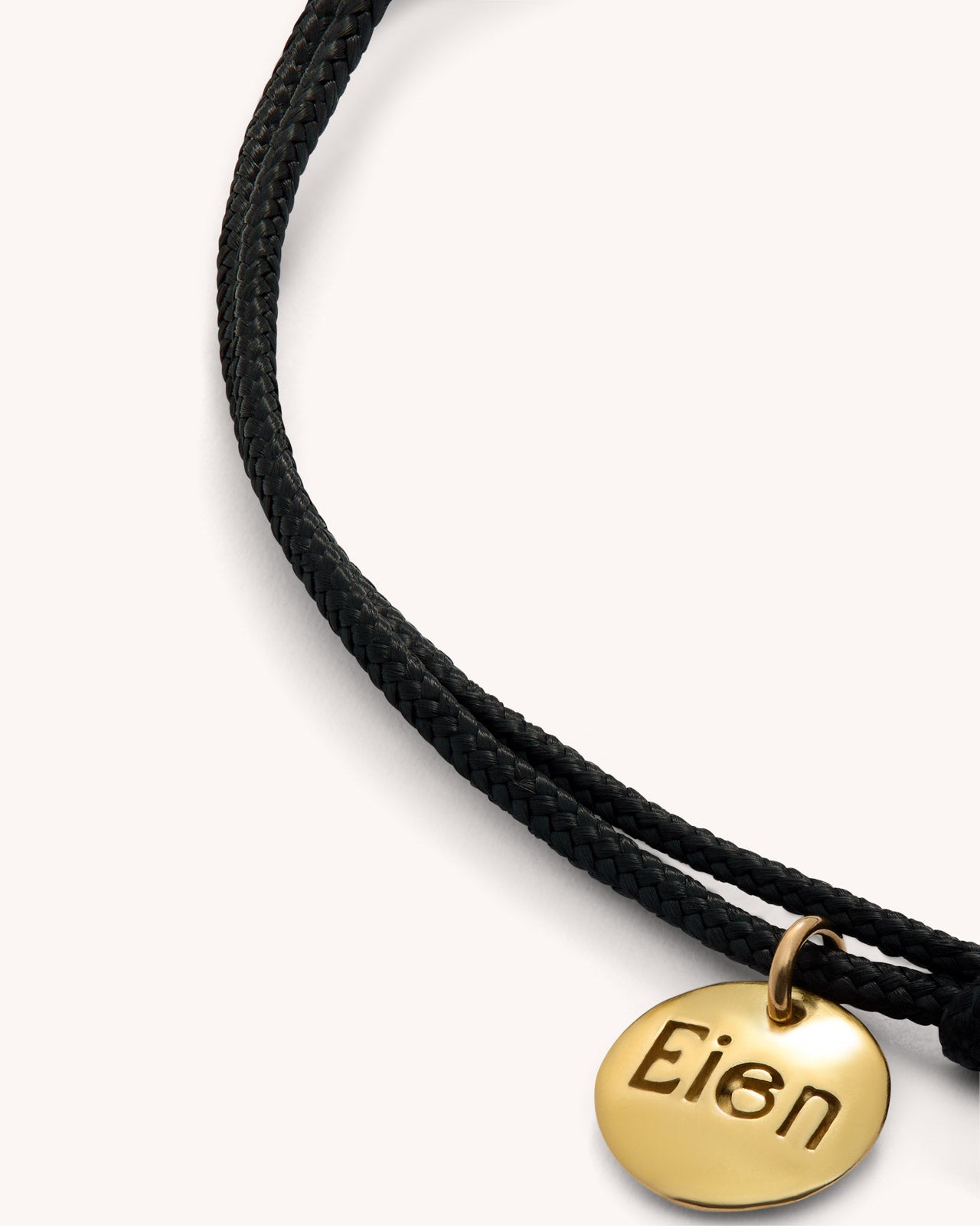 Detailed view of EIEN bracelet with engraved gold tag and double black cord, jewelry product photography.